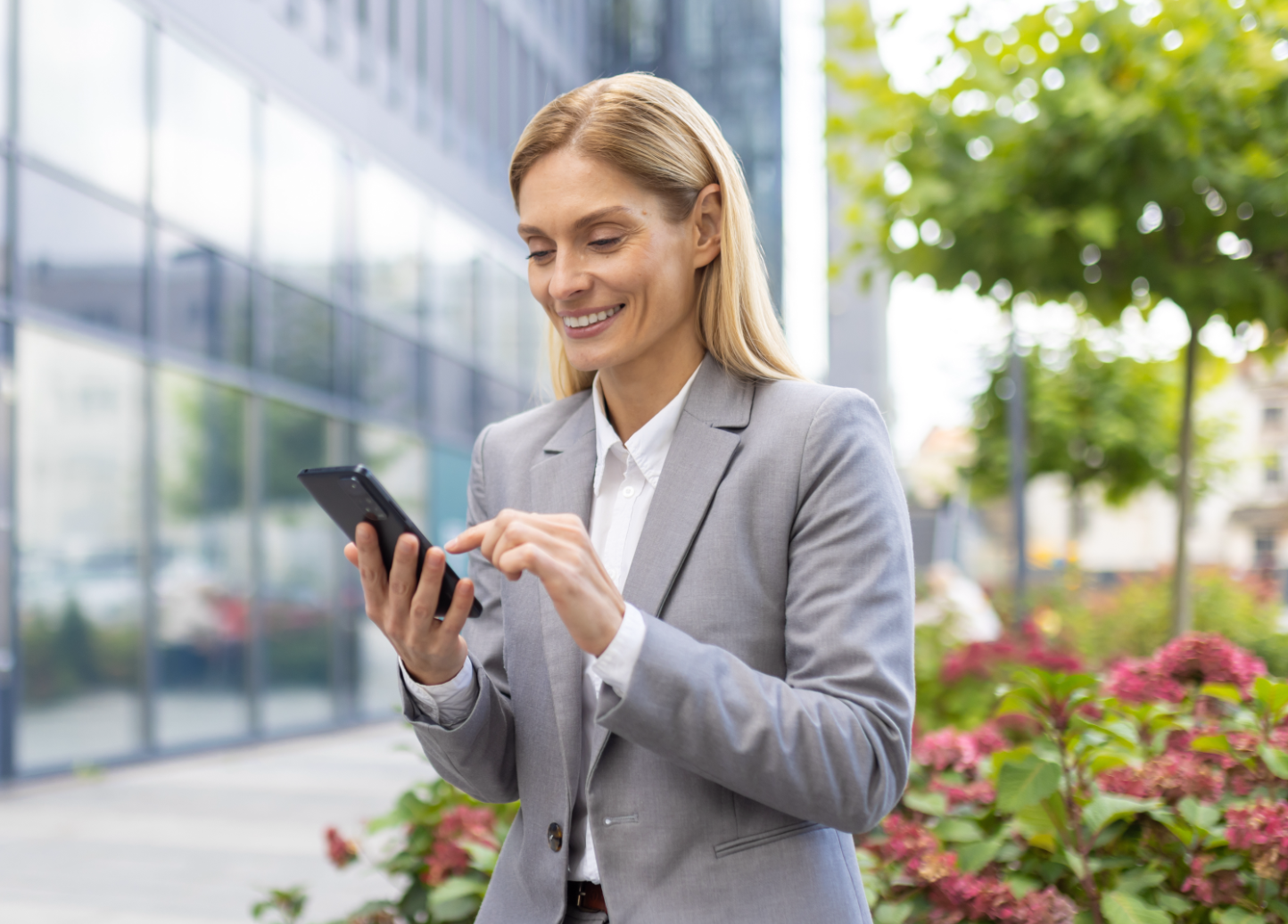 Businesswoman in gray suit using smartphone outdoors in modern office complex.